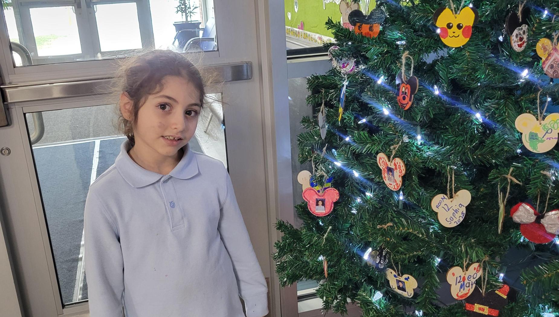 Young girl in a light blue shirt stands beside a decorated Christmas tree.