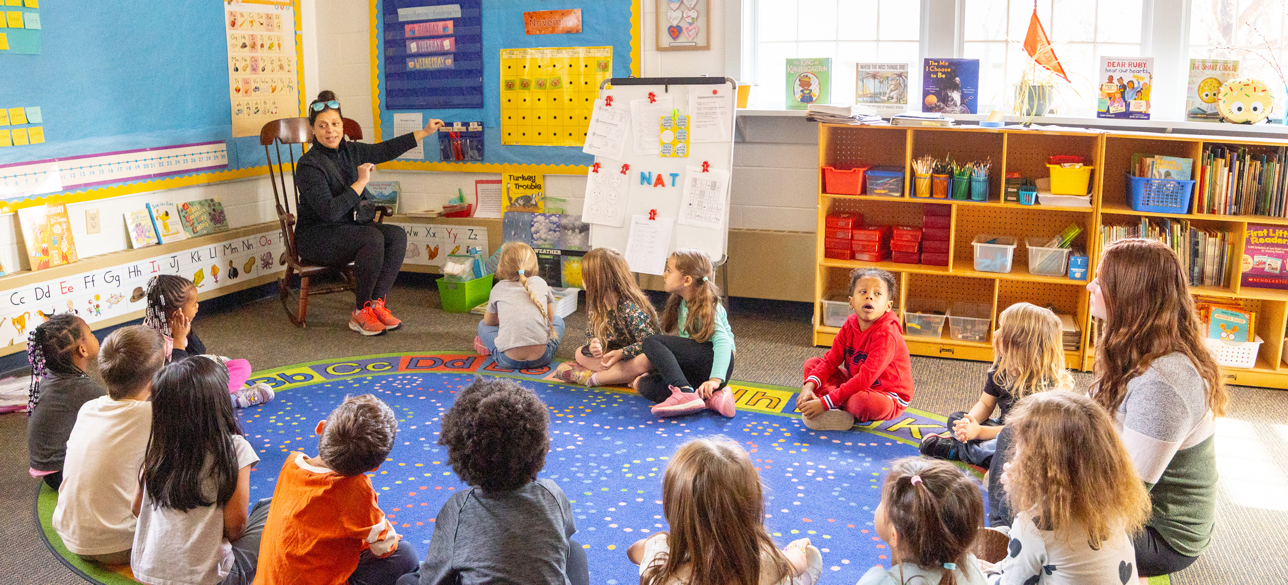 A teacher sits in a rocking chair leading a lesson for young students sitting in a circle on an alphabet rug inside a colourful classroom.