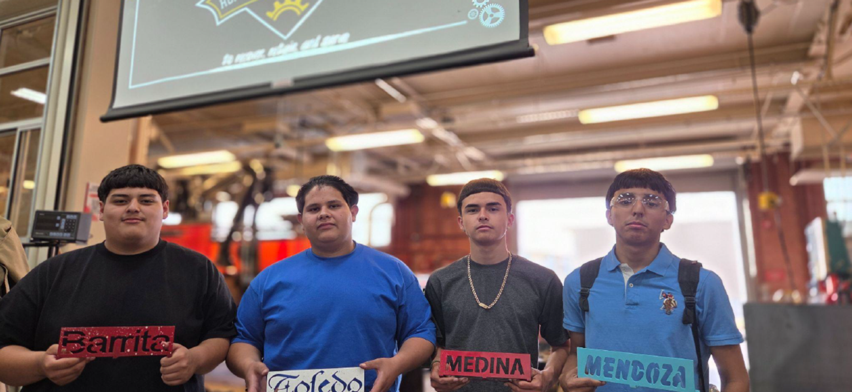 Four students hold signs with their names in an industrial workshop.