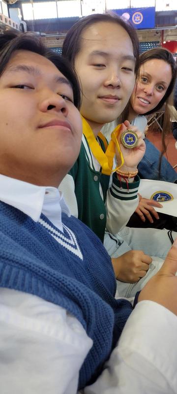 Two students show medals while seated with a third person nearby.