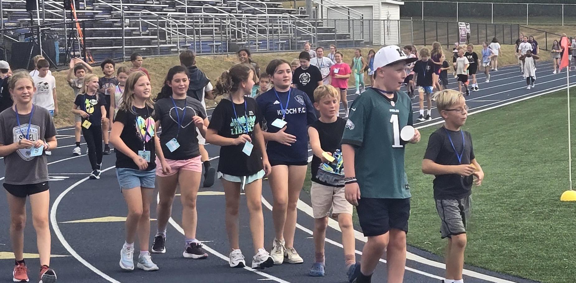 A group of children walking on a track, some holding cups and wearing lanyards.
