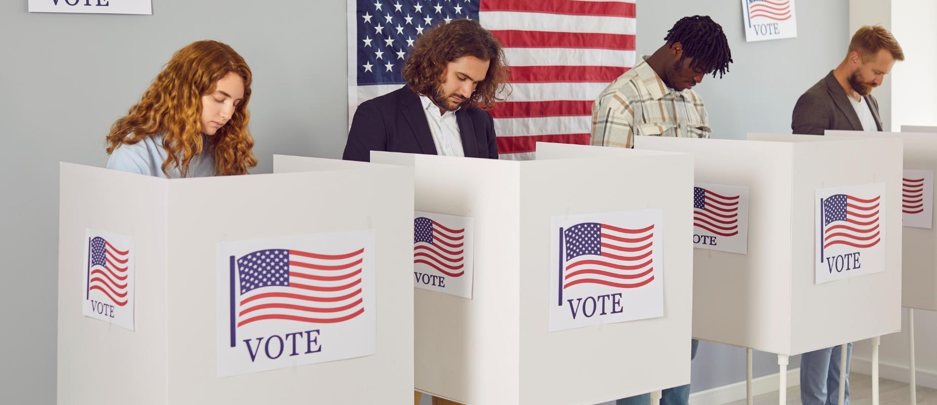 four adults vote behind partitions with the American flag on each partition as well as behind them