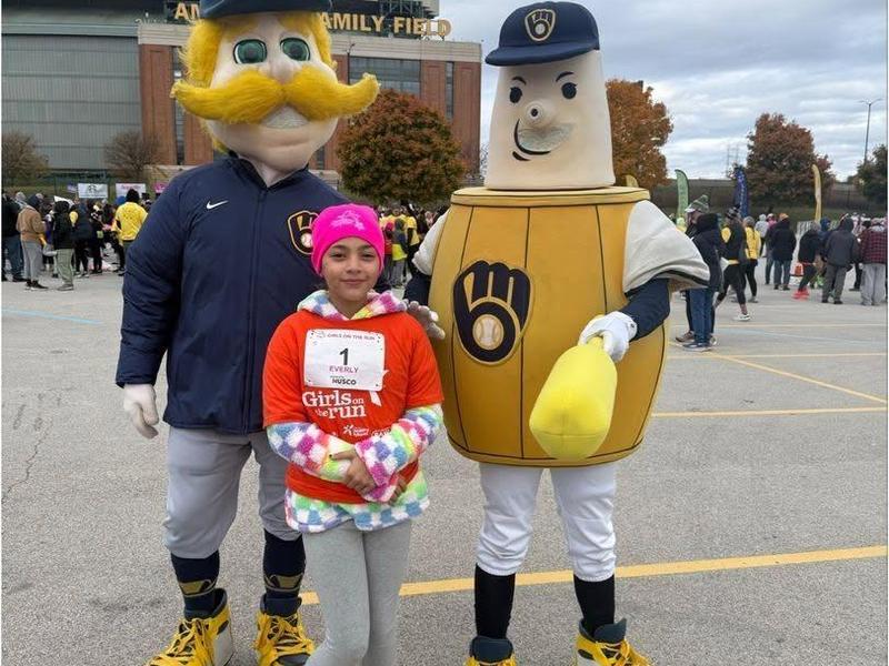 A girl standing between two mascots, one with a mustache and one resembling a beer barrel.