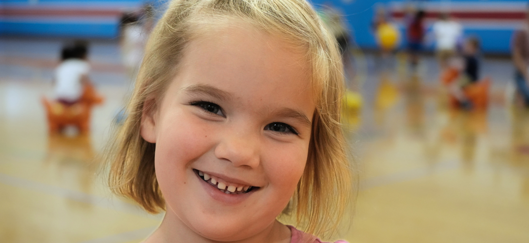 Smiling girl in a gym with children playing in the background.