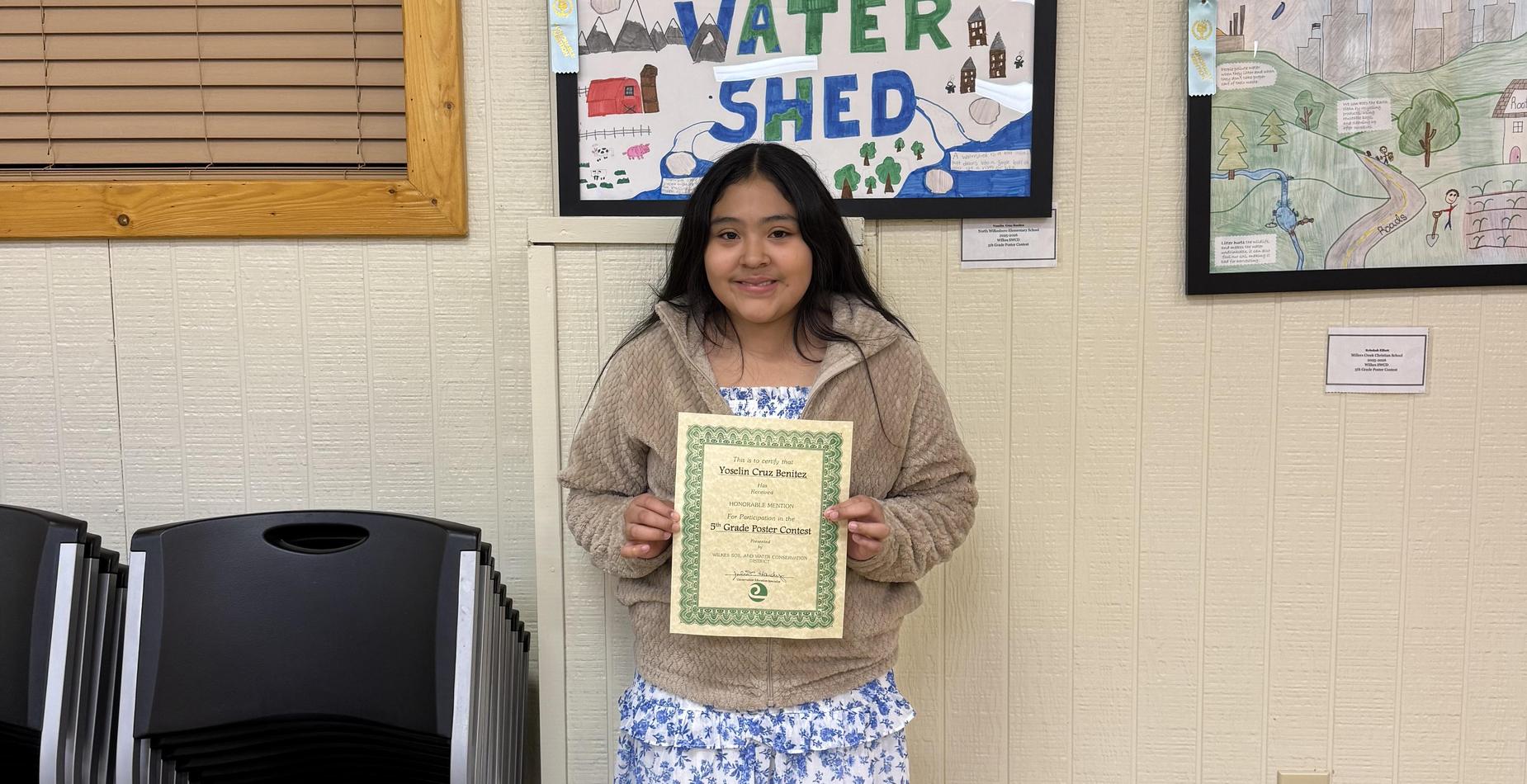 Child holding an award certificate in front of a colorful poster on the wall.