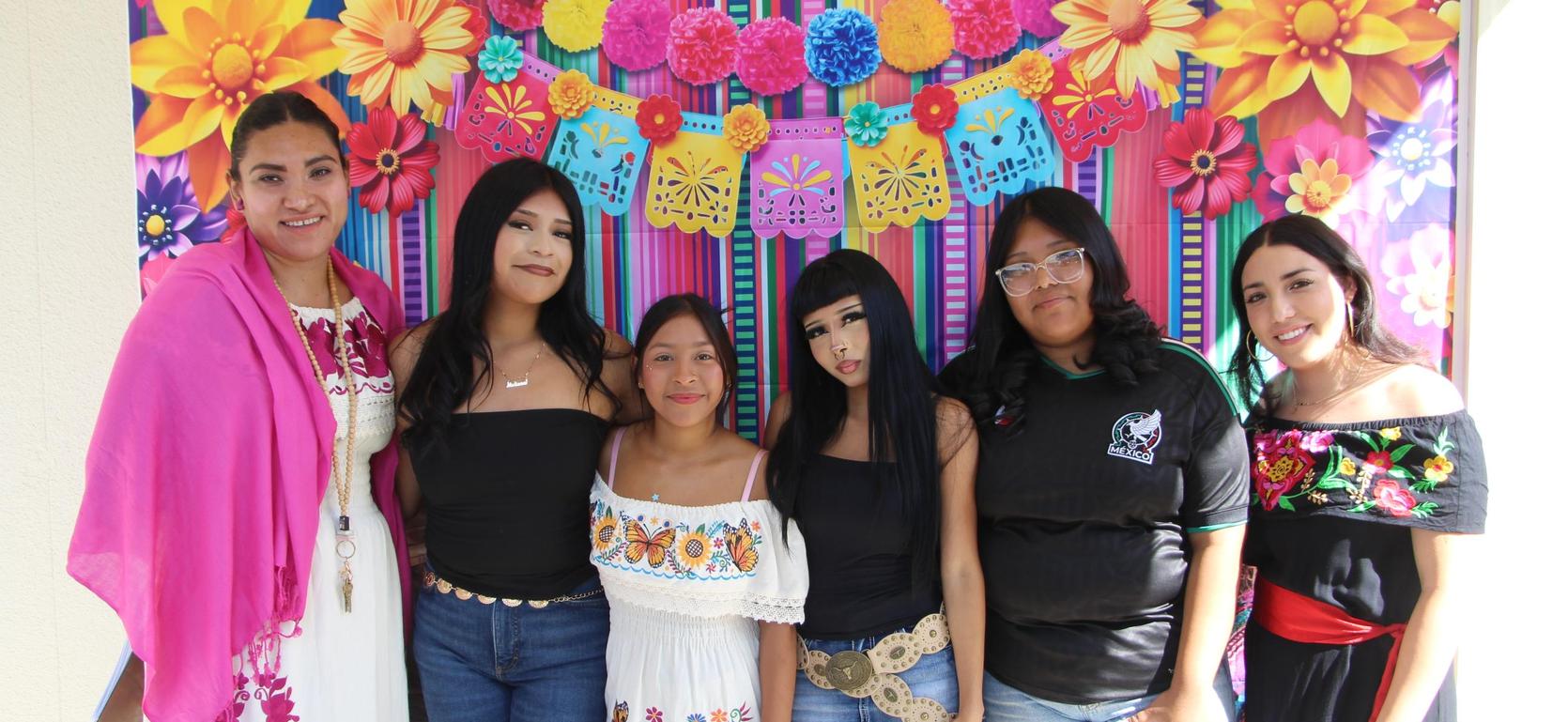 Group of six women in colorful outfits posing against a vibrant floral backdrop.
