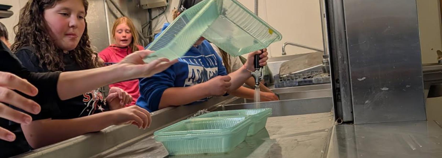 Children washing plastic containers at a sink in a kitchen setting.