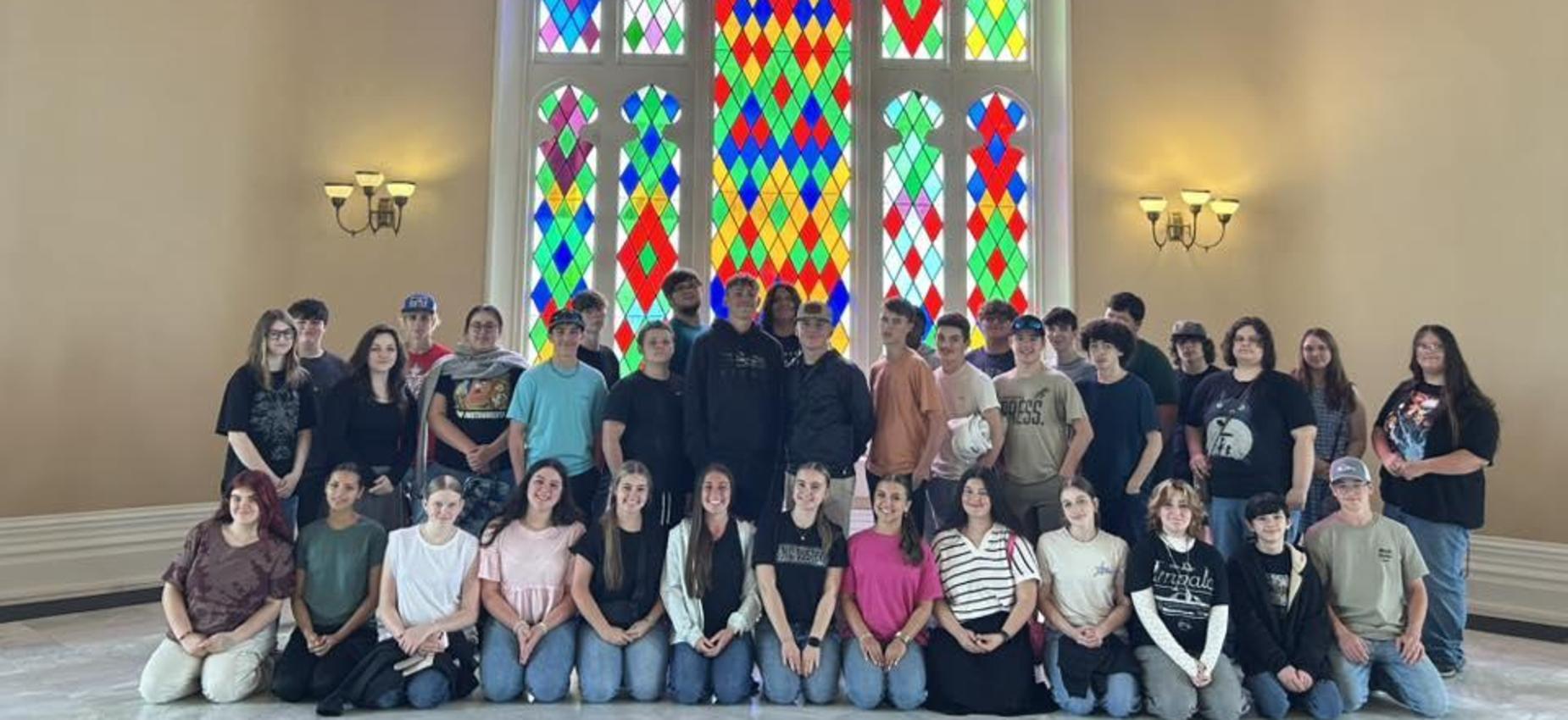 A large group of young people posing in front of stained glass windows.
