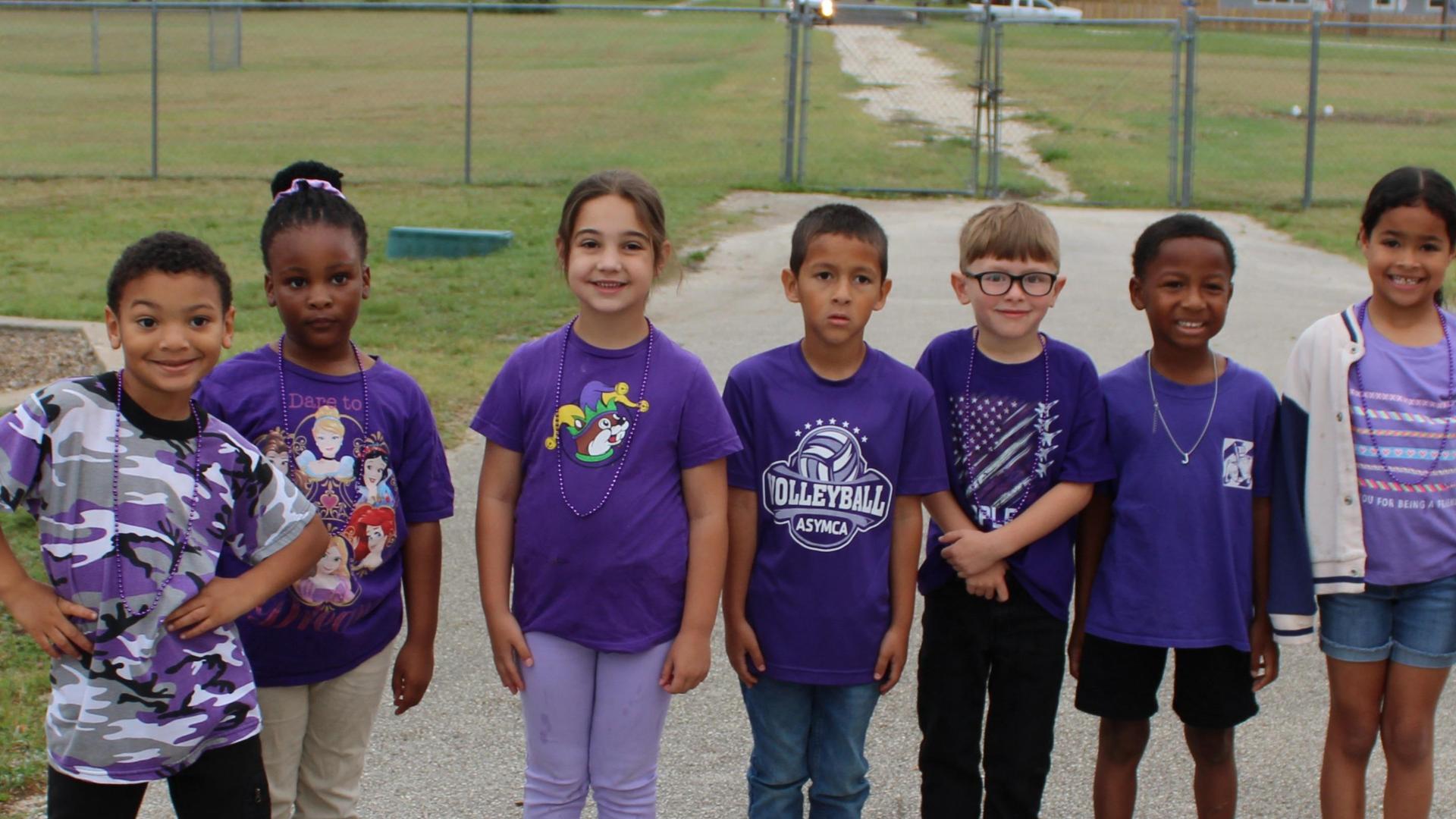 Group of seven children wearing purple shirts posing outside.