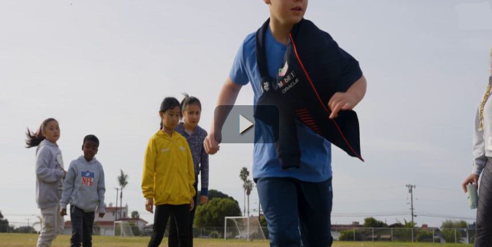 A boy running on a field with several kids in the background watching.