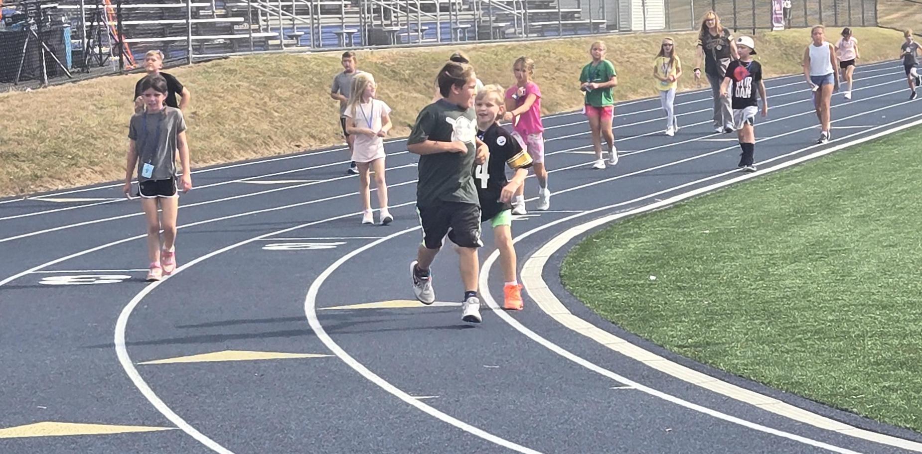 Children jogging on a track, some wearing sports attire and medals.