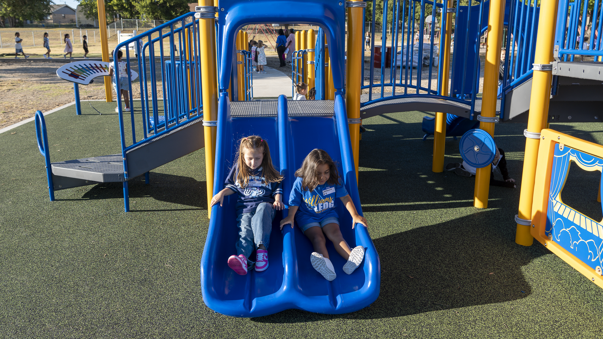 Two children sliding down a blue playground slide on a sunny day.