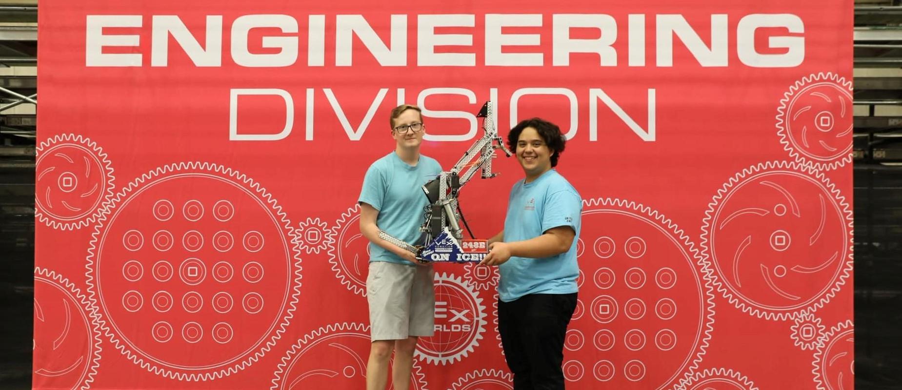two young men with robot in hand in front of engineering division sign