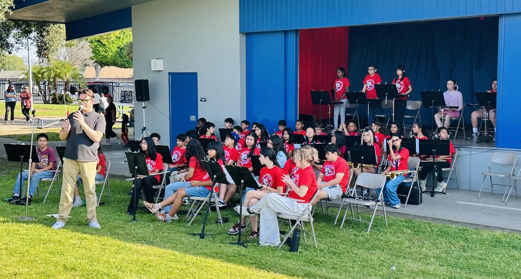Band performing outdoors on stage with a conductor in front.