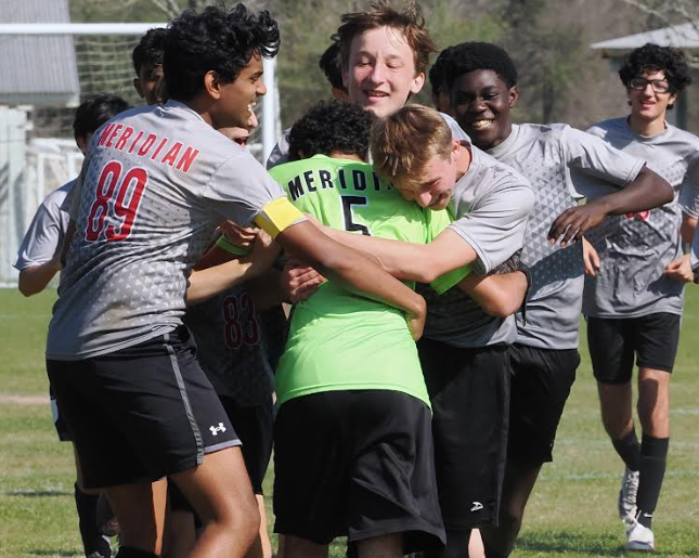 Students in soccer uniforms run towards their goalie, smiling