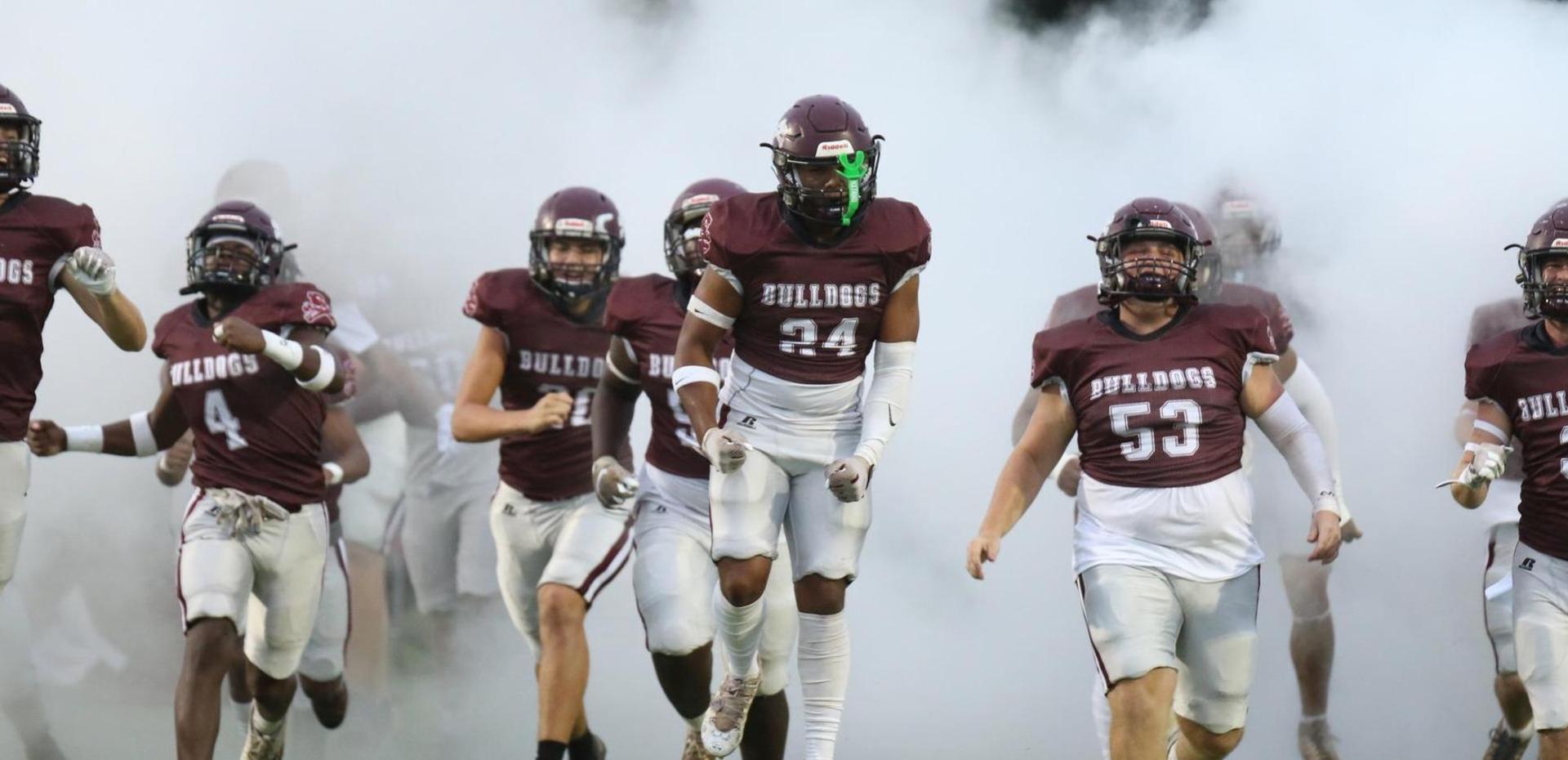 Football players in maroon uniforms running through smoke onto the field.