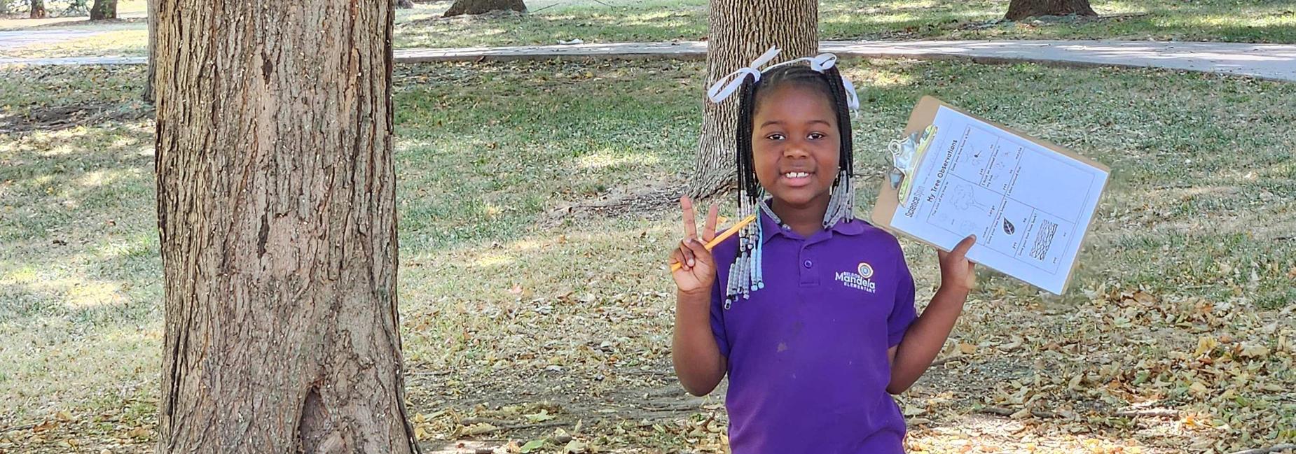 A scholar stands outside with a clipboard and poses for a picture with a peace sign