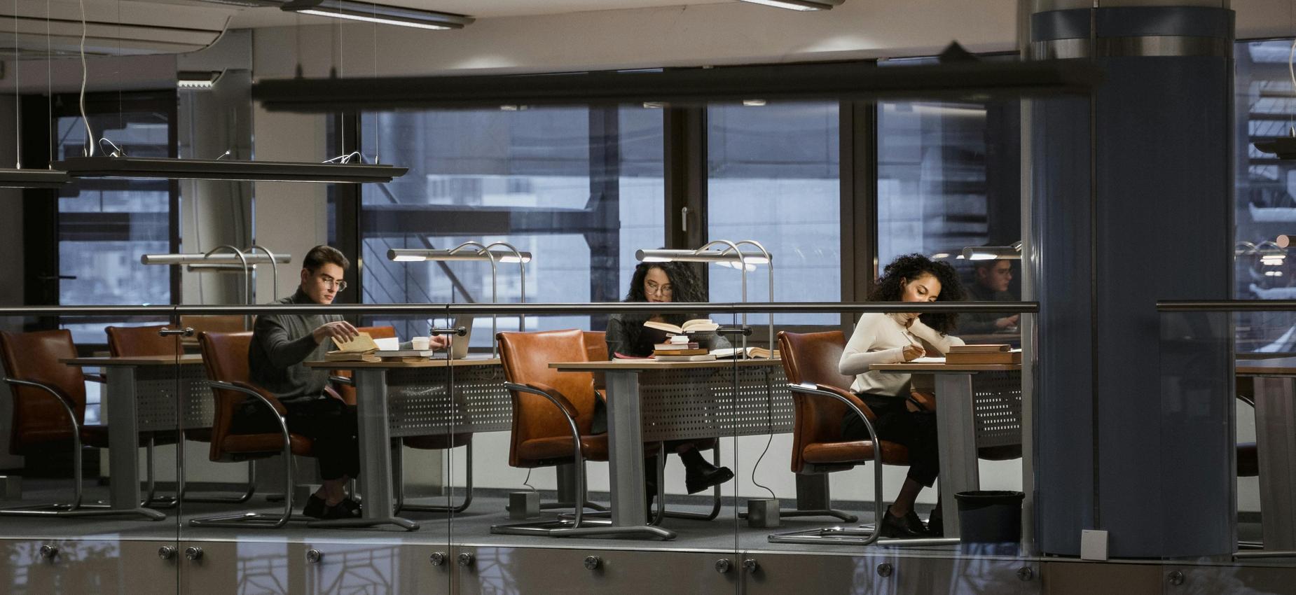 Three individuals reading books in a modern library setting.