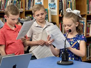 A young girl reads and speaks into a mic as two other students listen