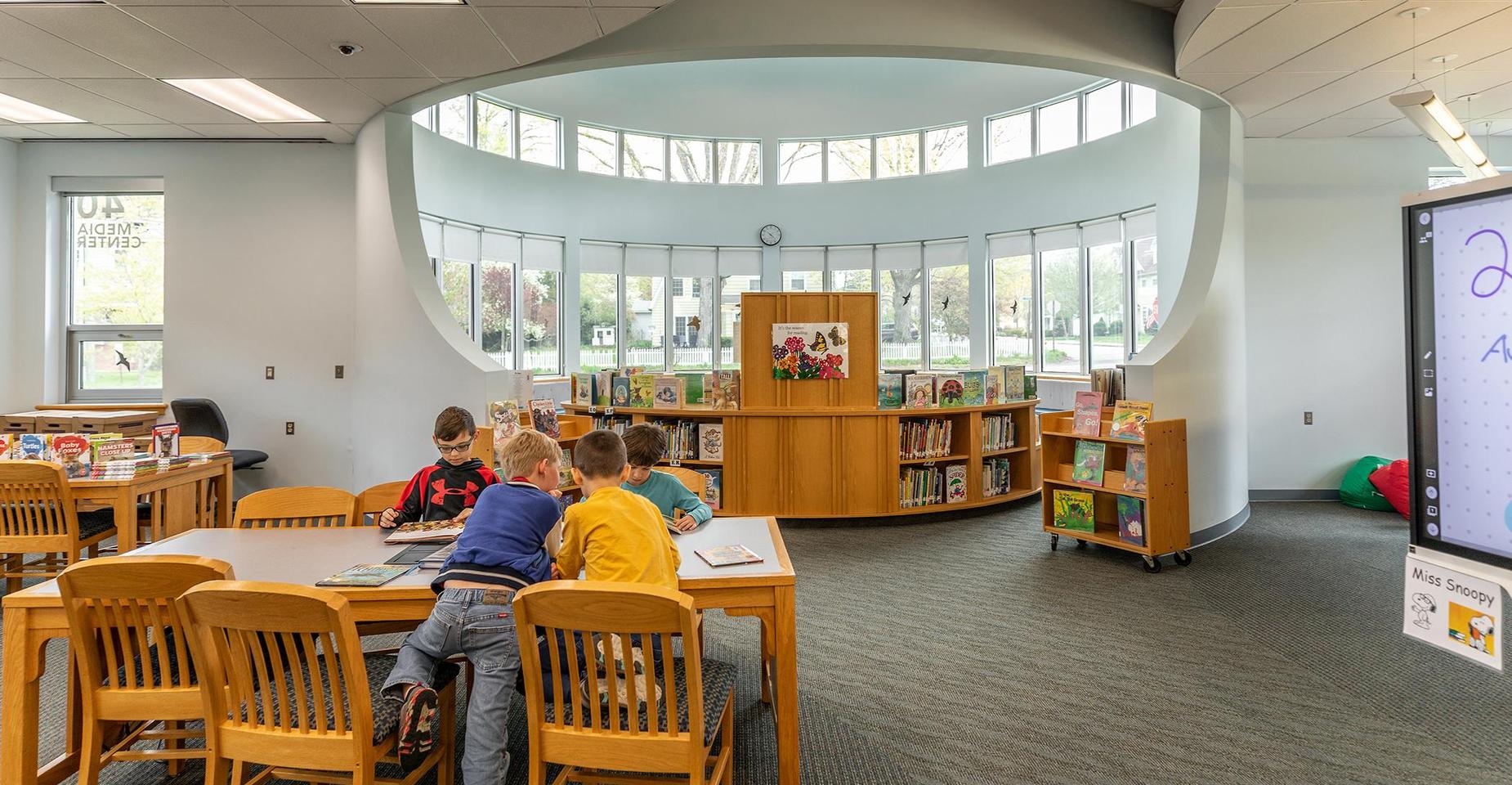 Children sitting at tables in a library with shelves and large windows.
