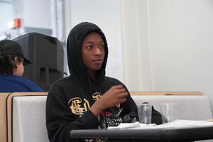 Young person in a hoodie sitting at a table, looking thoughtful.