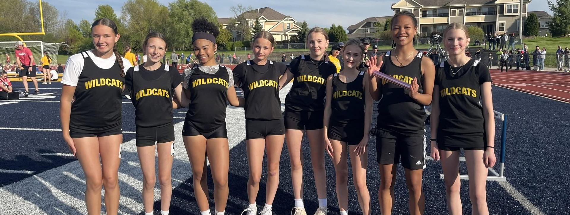 A group of eight girls in track uniforms smiling together on a field.