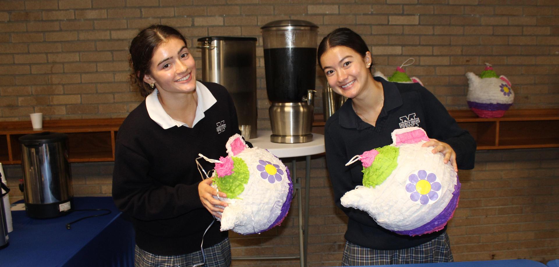 Two Mercy Students on Mercy Day holding colorful teapot-shaped pi&ntilde;atas with beverages on a table.