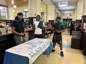 Three students pose by a table with a signed banner in a lively library setting.