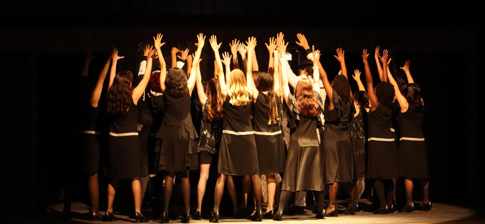 A group of women in black dresses stand with hands raised in a theatrical setting.