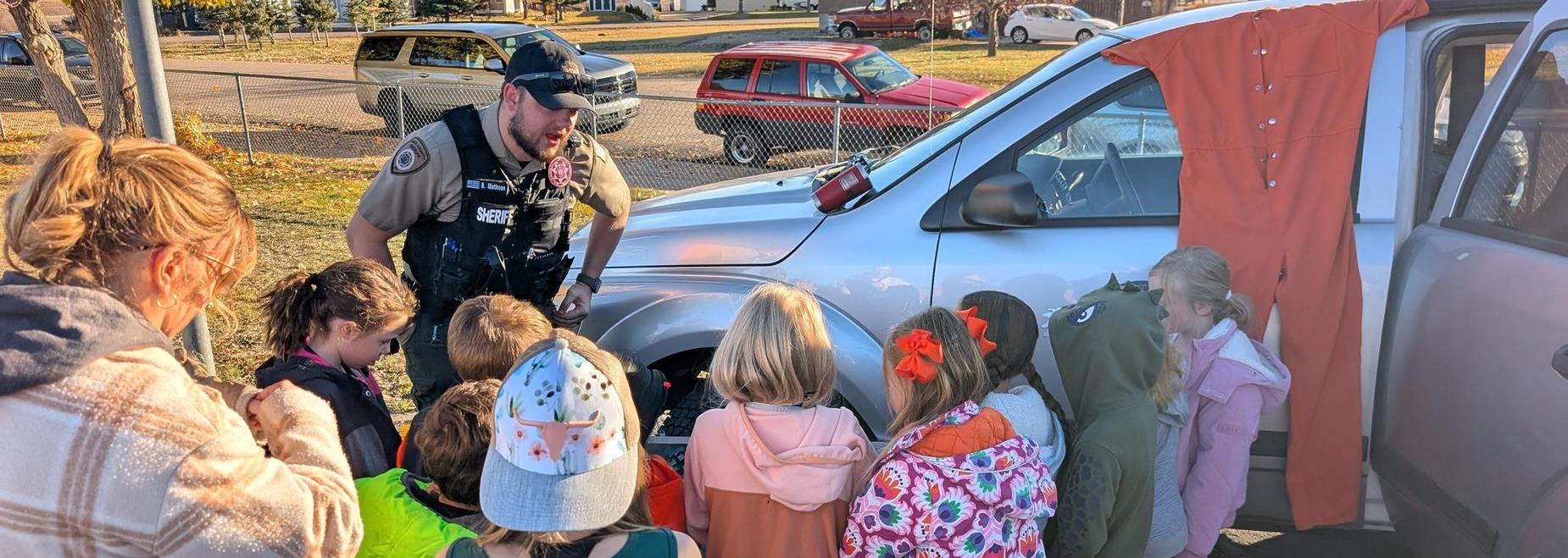 Children gathered around a law enforcement officer by a vehicle, learning about safety.