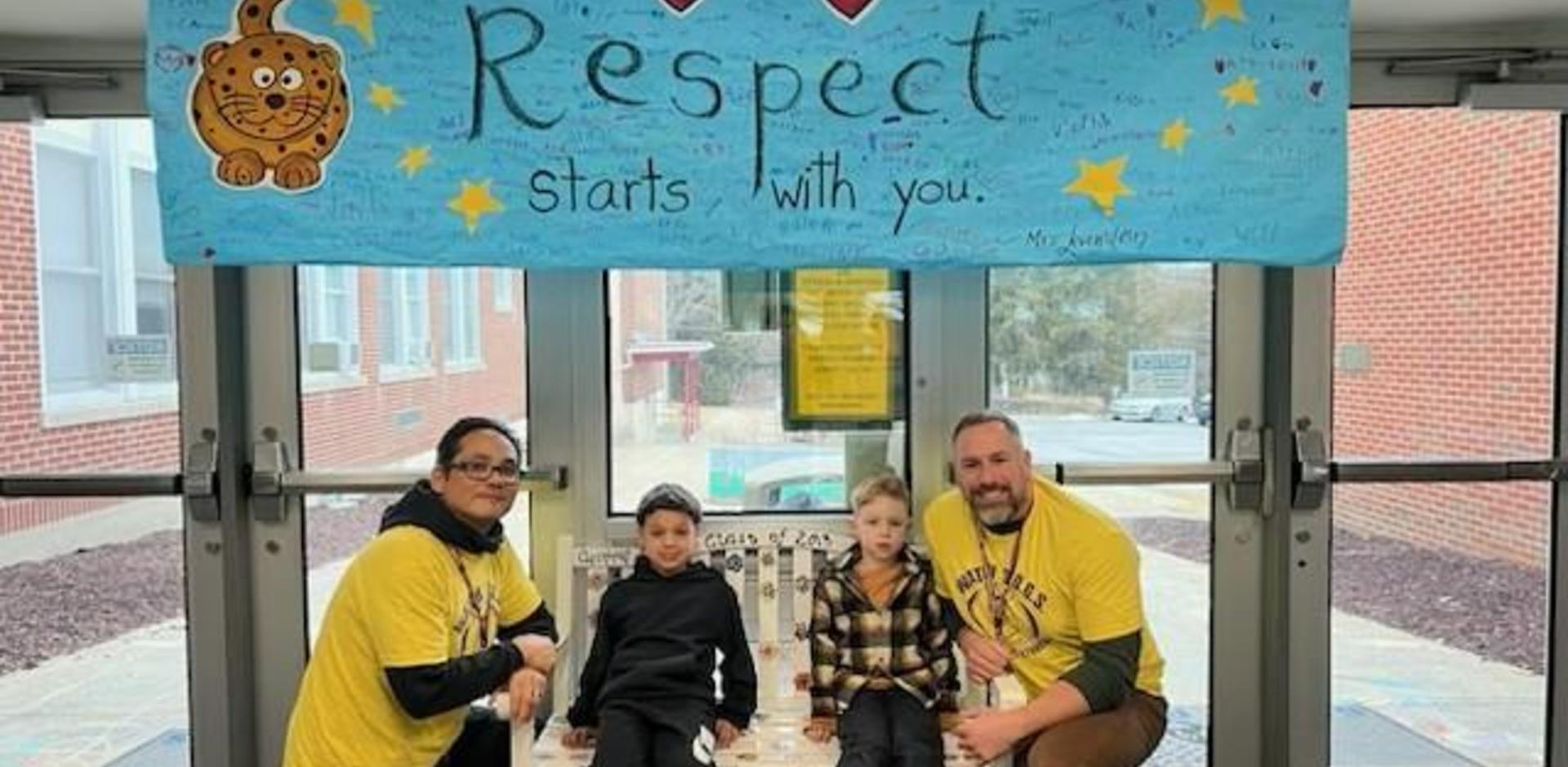 Two adults and two children seated under a 'Respect starts with you' banner at a school entrance.