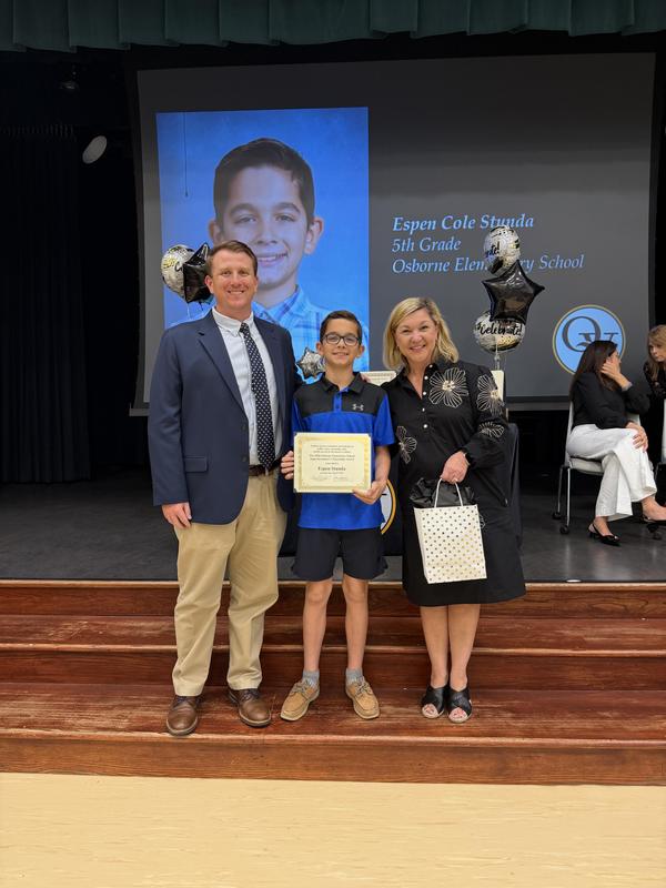 Student holding a certificate is standing between his principal and superintendent on a stage.