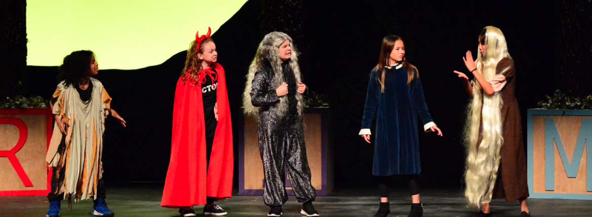 Texas School for the Deaf (TSD) middle school students perform on stage during The Brothers Grimm Spectaculathon. The scene features five students in colorful costumes, including a tattered outfit, a devil's cape and horns, a wolf costume, a blue velvet dress, and a character with long, flowing hair, engaging in an animated and expressive dialogue under a dramatic spotlight.