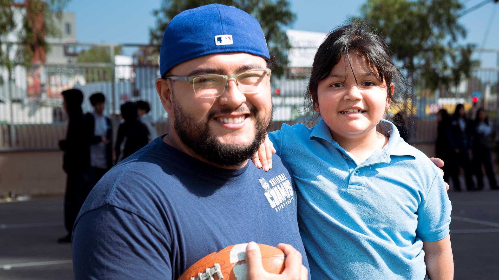 A man holding a football stands next to a smiling child outdoors.