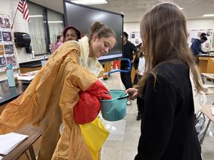 Student in a colorful poncho handing items to a classmate in a lab setting.