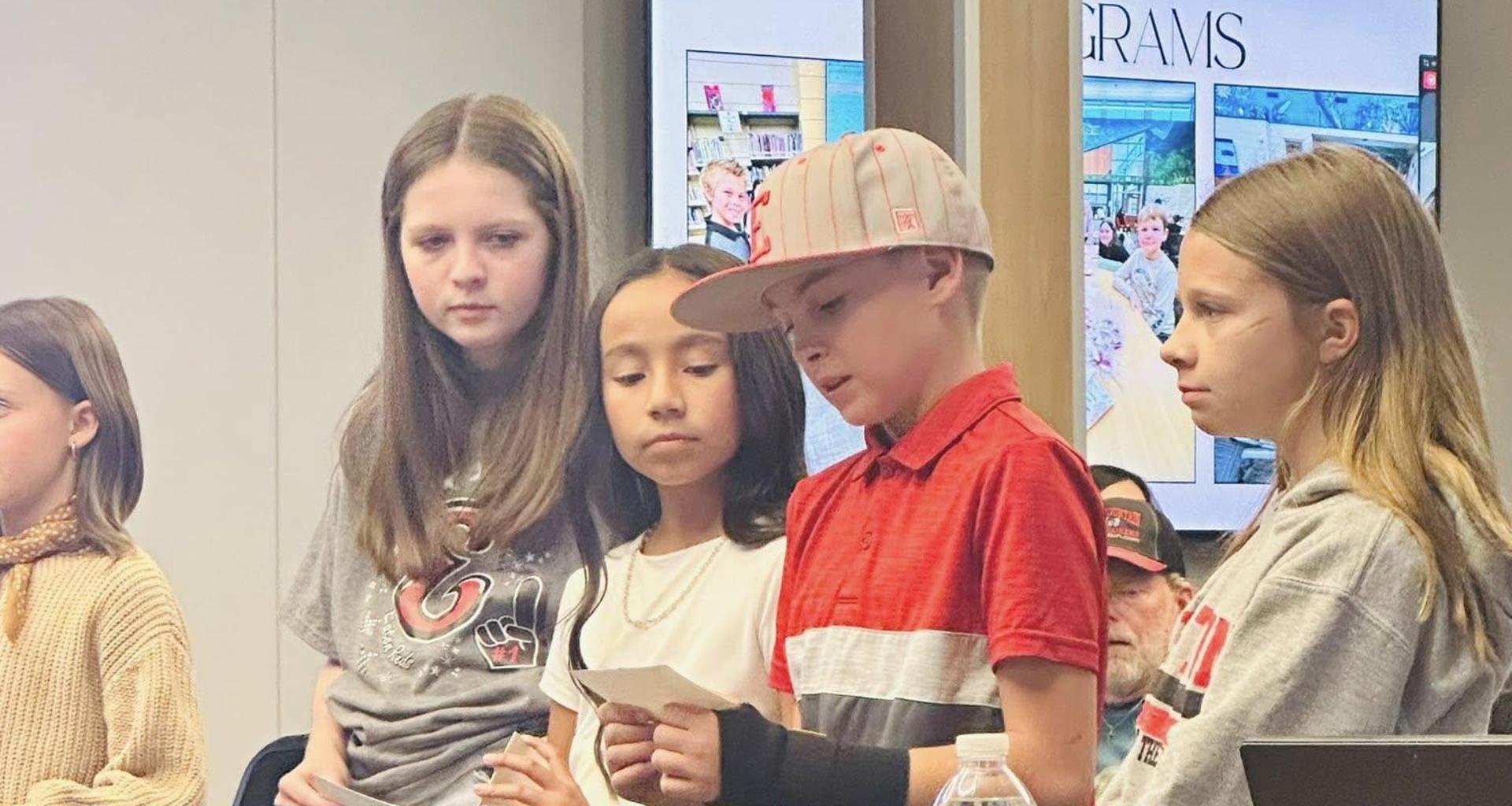 Students presenting at a table, holding papers and listening attentively.