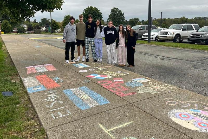 Group of six students posing beside colorful chalk drawings on a sidewalk.