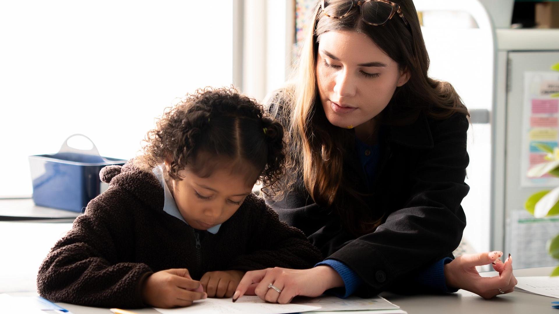 A woman helping a girl with her writing at a table.