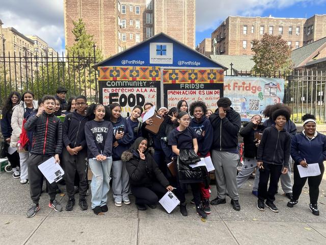 Large group of students posing in front of a community fridge mural with smiles.