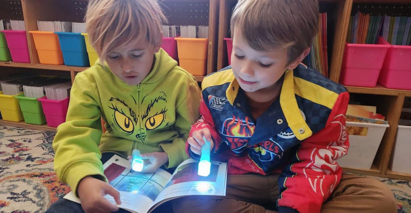 Two children reading a book while using small light devices to illuminate the pages.