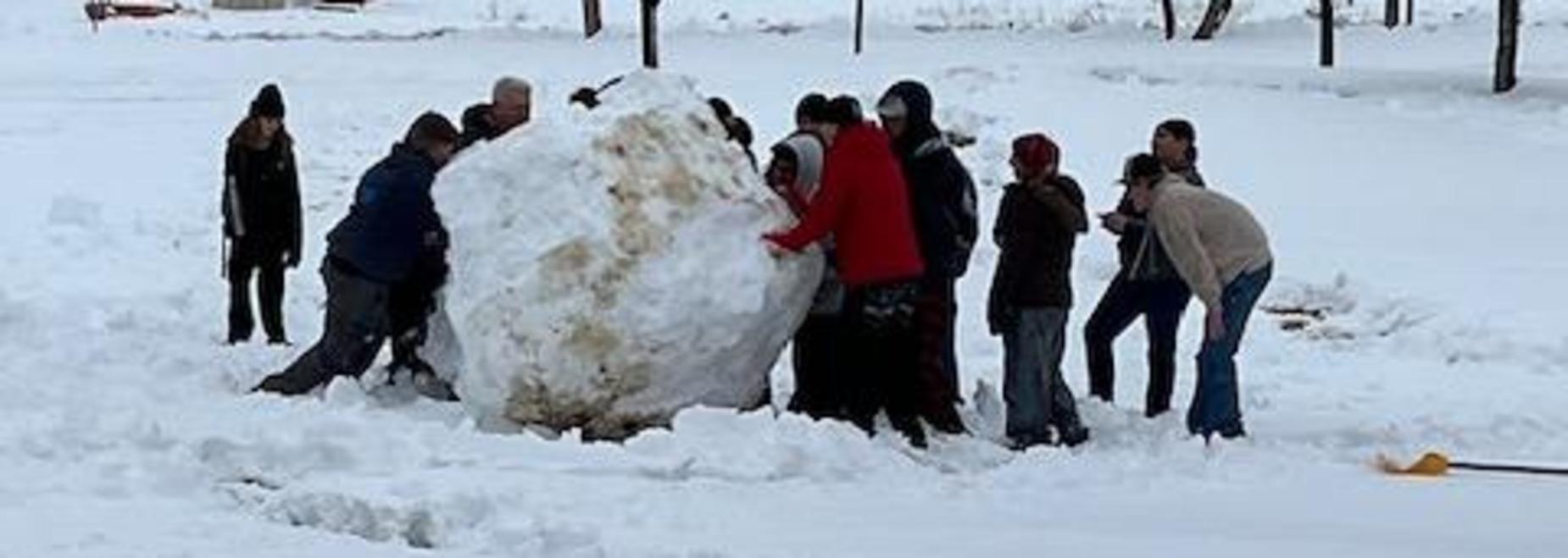 Different angle of the same group moving a large snowball in a snowy area.