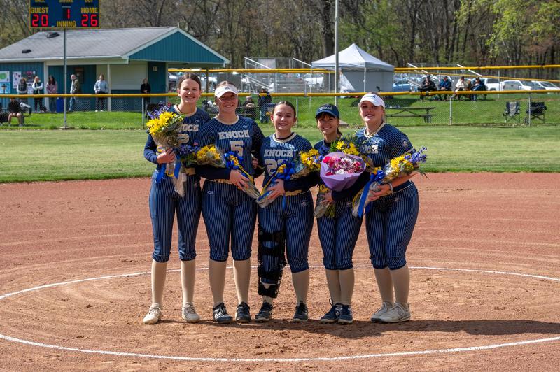 5 softball players dressed in uniforms on field holding flowers