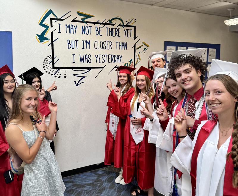 Students in graduation caps and gowns point toward an inspirational quote mural on a school hallway wall.