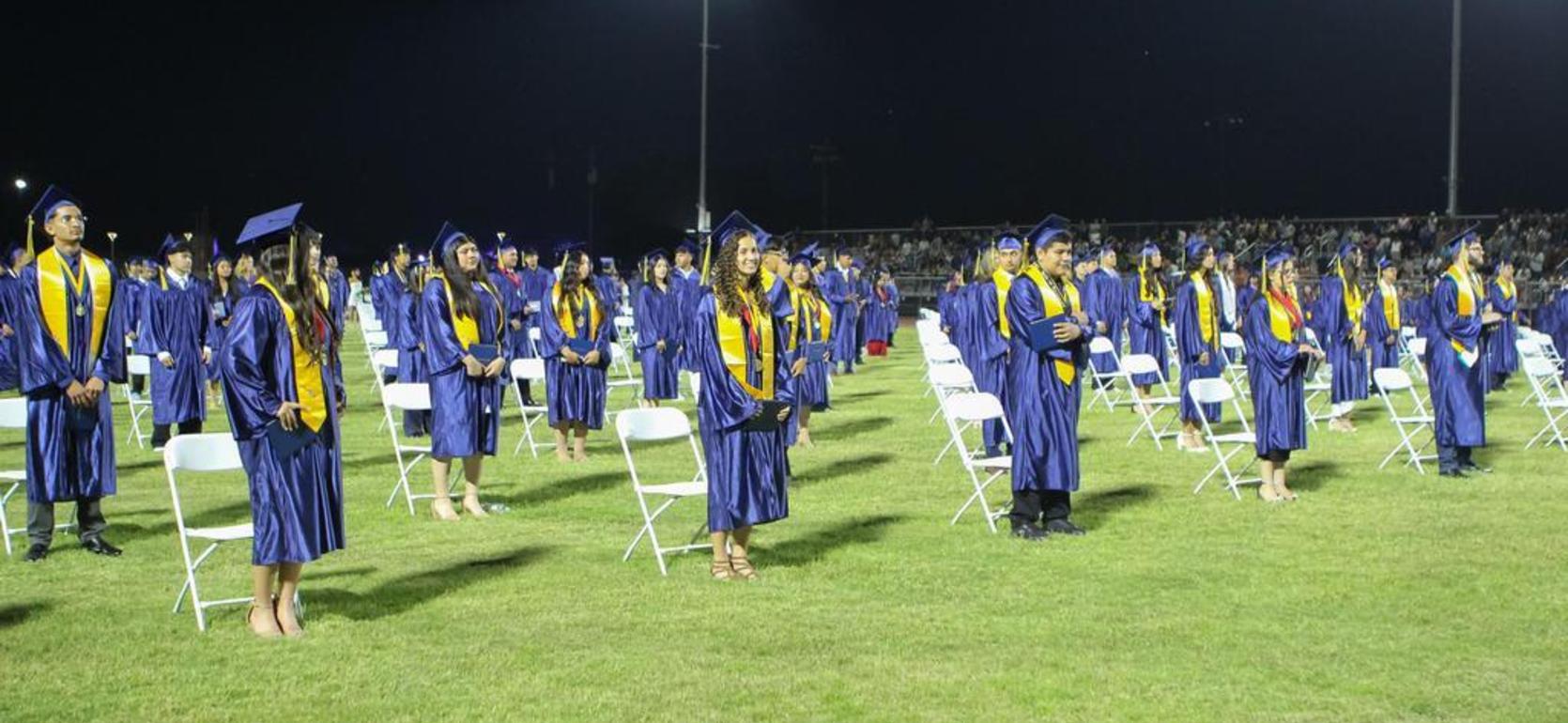 Graduation ceremony at night with graduates standing in rows on a field, wearing caps and gowns.