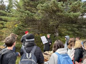 A group observes a presenter showing a poster in a forested area.