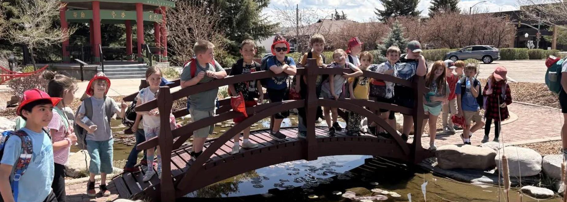 Group of children posing on a wooden bridge near a pond with greenery.