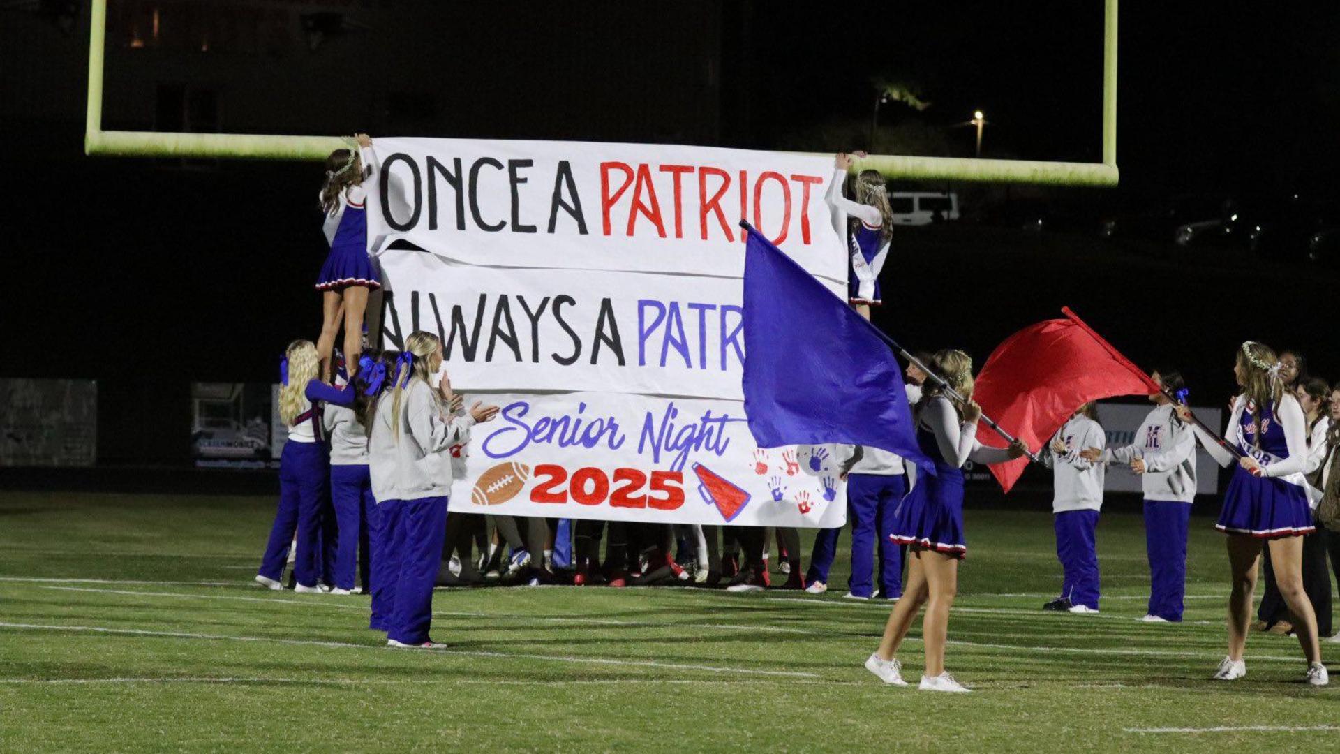 Cheerleaders hold a banner for Senior Night as others wave flags on the field.