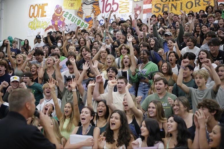 A crowd of students in the gymnasium bleachers cheer in excitement at a school rally.