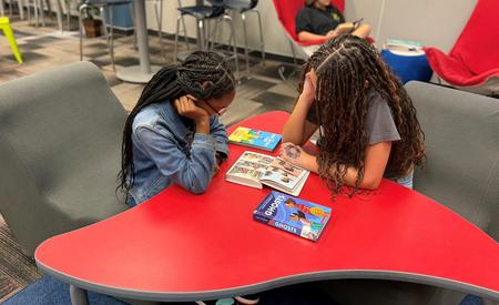 Two girls reading books at a red table in a classroom setting.