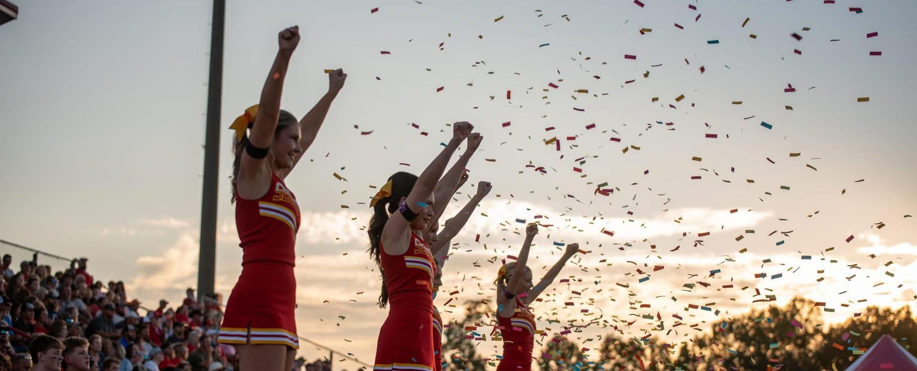 Cheerleaders in red uniforms celebrating with confetti and an audience in the background.